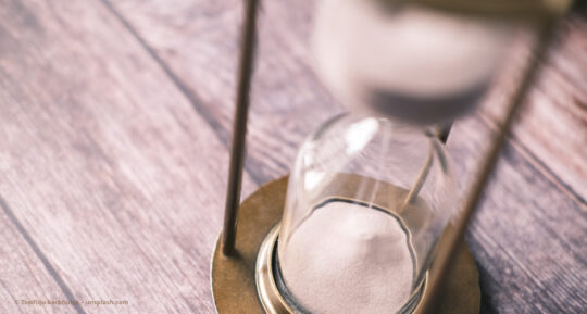 Eine Sanduhr steht auf Holzbalken. Der weiße Sand rinnt hinunter. © Towfiqu barbhuiya – unsplash.com